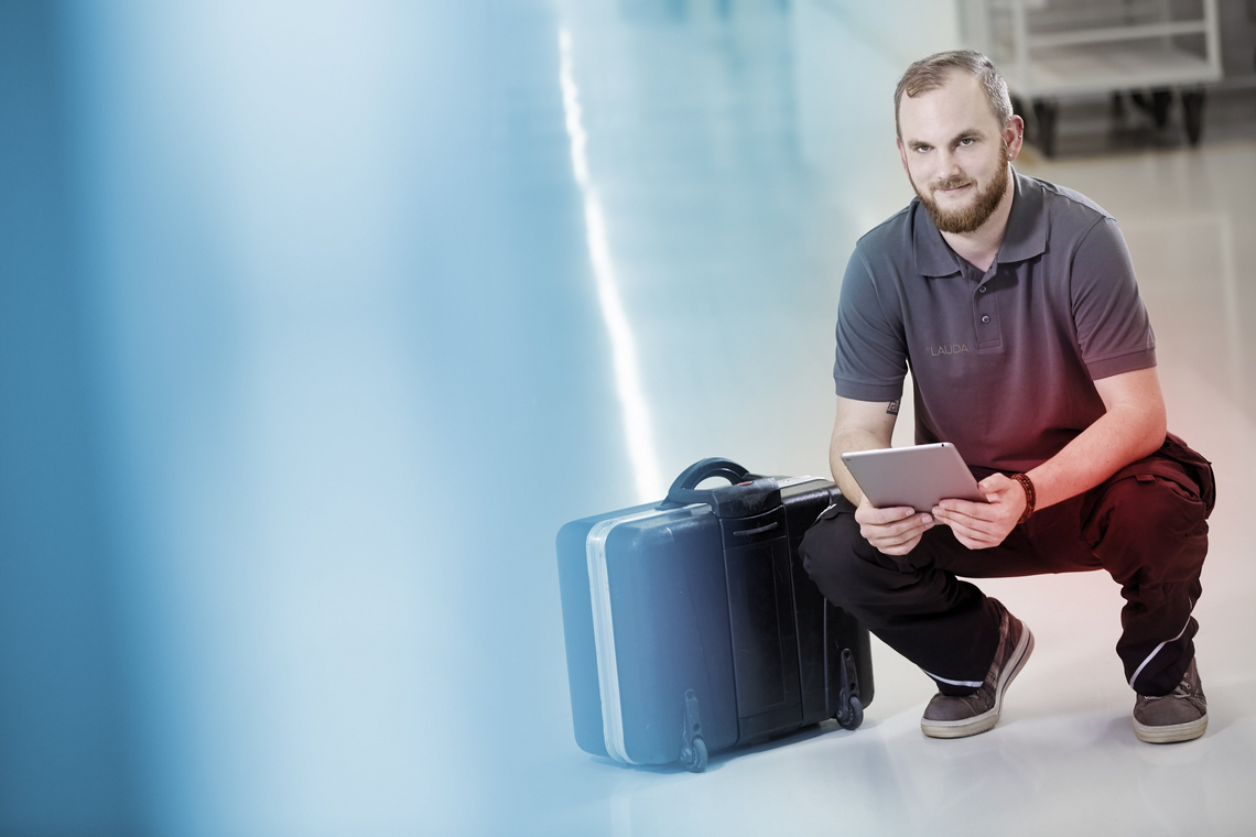 Сотрудник сервисной службы LAUDA стоит на коленях возле черного чемодана с планшетом в руке.